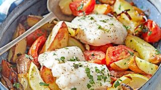 A baked dish perfect for Spring One Pot Dinners&mdash;white fish fillets, roasted potato wedges, tomatoes, lemon slices, and fresh herbs in a blue baking dish with a metal serving spoon.