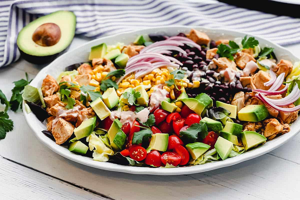 A large chicken taco salad on a white plate with chopped avocado, tomatoes, corn, black beans, red onion, lettuce, cilantro, and dressing. Half an avocado sits beside the plate.