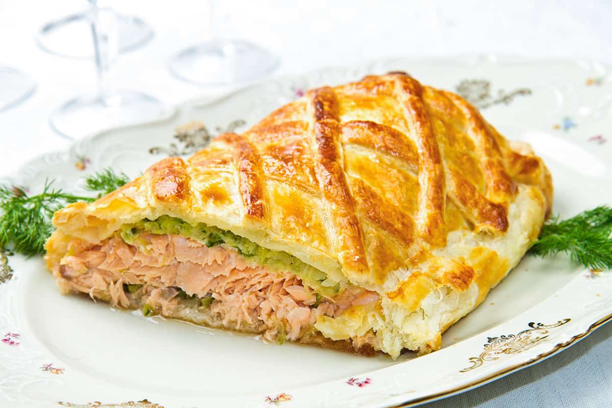 A slice of Salmon Wellington with golden puff pastry, filled with tender salmon and a layer of spinach, served on a decorated plate with fresh dill garnish.