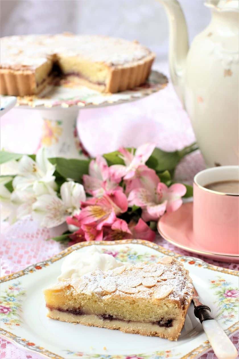 A slice of almond tart with a layer of jam, served with whipped cream on a floral plate, alongside coffee, flowers, and a teapot-perfect for an elegant Downton Abbey Afternoon Tea.