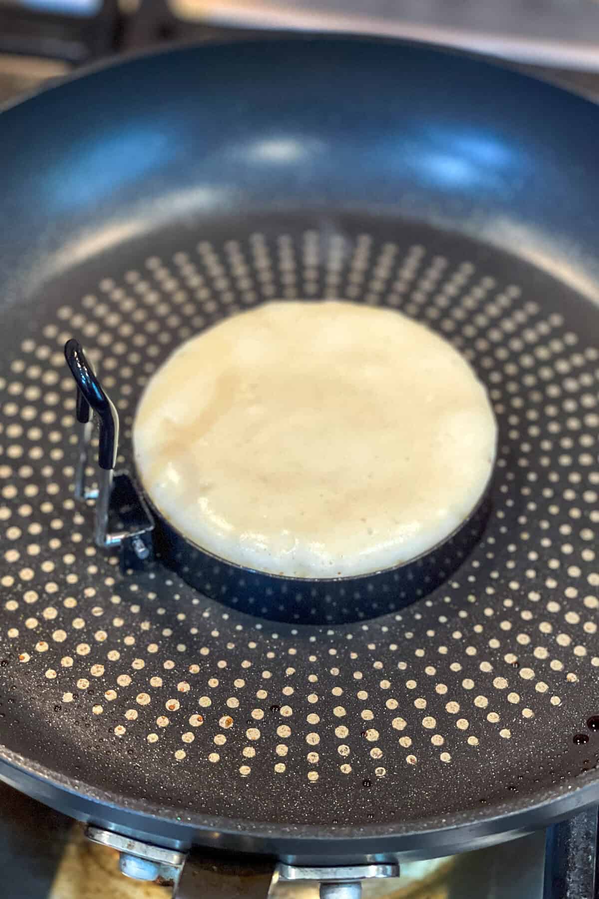 A round pancake batter from a fluffy pancake recipe is cooking in a non-stick frying pan, held in shape by a circular metal mold. The pan features a dotted pattern on its surface.