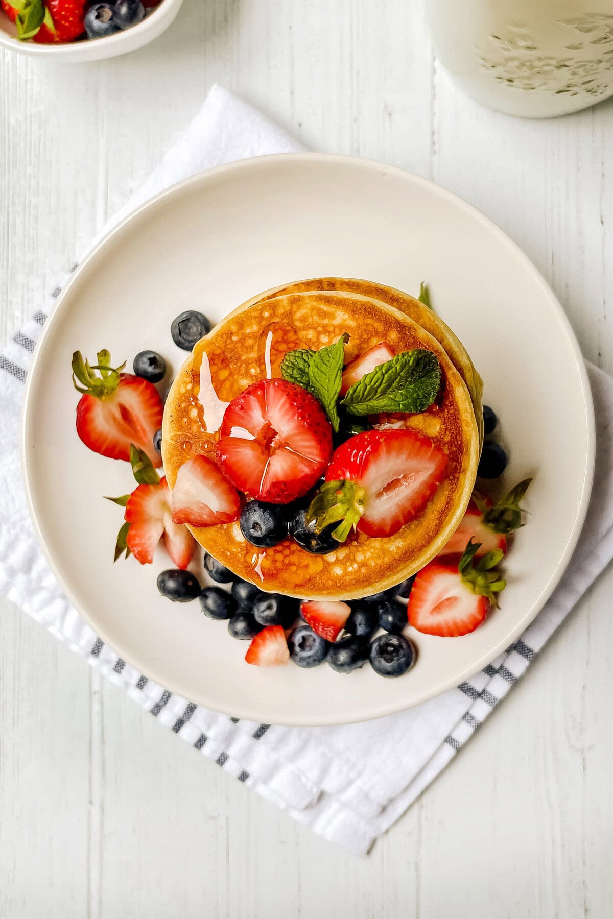 A stack of pancakes from a fluffy pancake recipe is topped with sliced strawberries, blueberries, and mint leaves on a white plate. Extra berries are scattered around the dish, which sits on a striped napkin atop a white wooden surface.