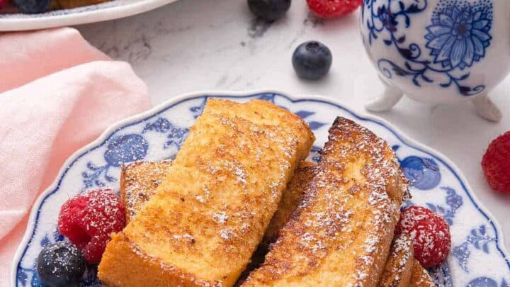 Plates of French toast topped with powdered sugar, raspberries, and blueberries, next to a small jug of syrup on a white surface—perfect for Mother’s Day brunch ideas.