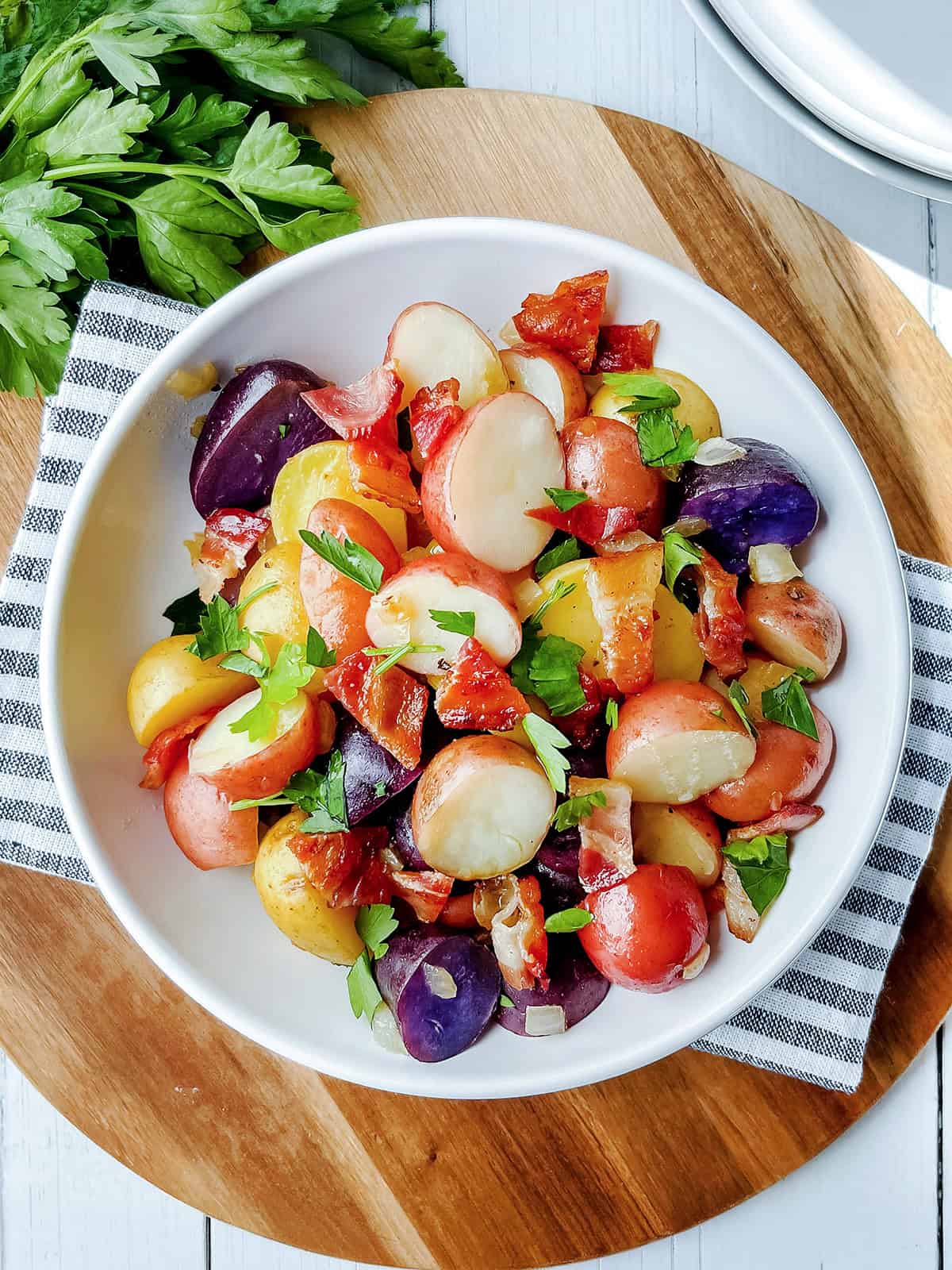 A bowl of German Potato Salad featuring red, yellow, and purple potatoes, chopped parsley, and pieces of bacon on a wooden board with a striped cloth and fresh parsley in the background.
