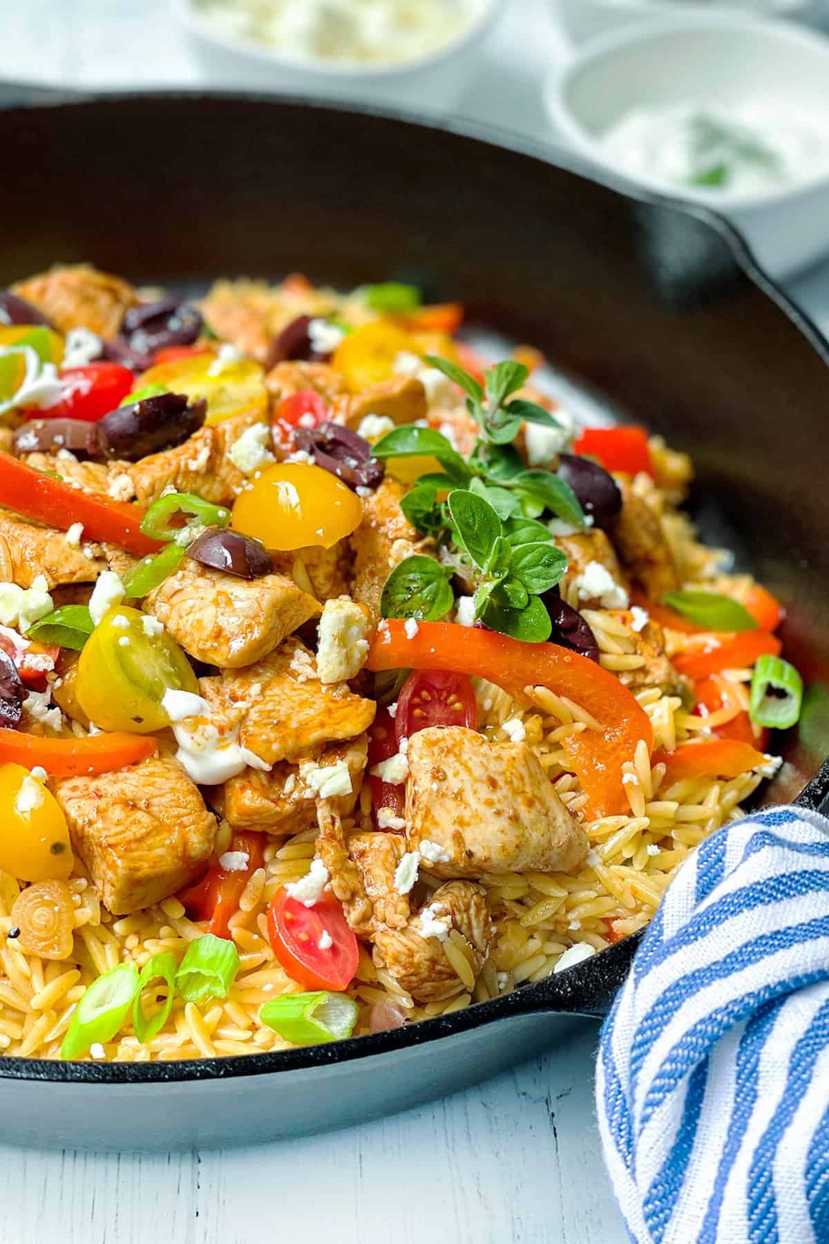 A black skillet filled with skillet Greek chicken, cooked orzo, sliced bell peppers, cherry tomatoes, olives, feta cheese, and fresh herbs, with a striped towel on the side.