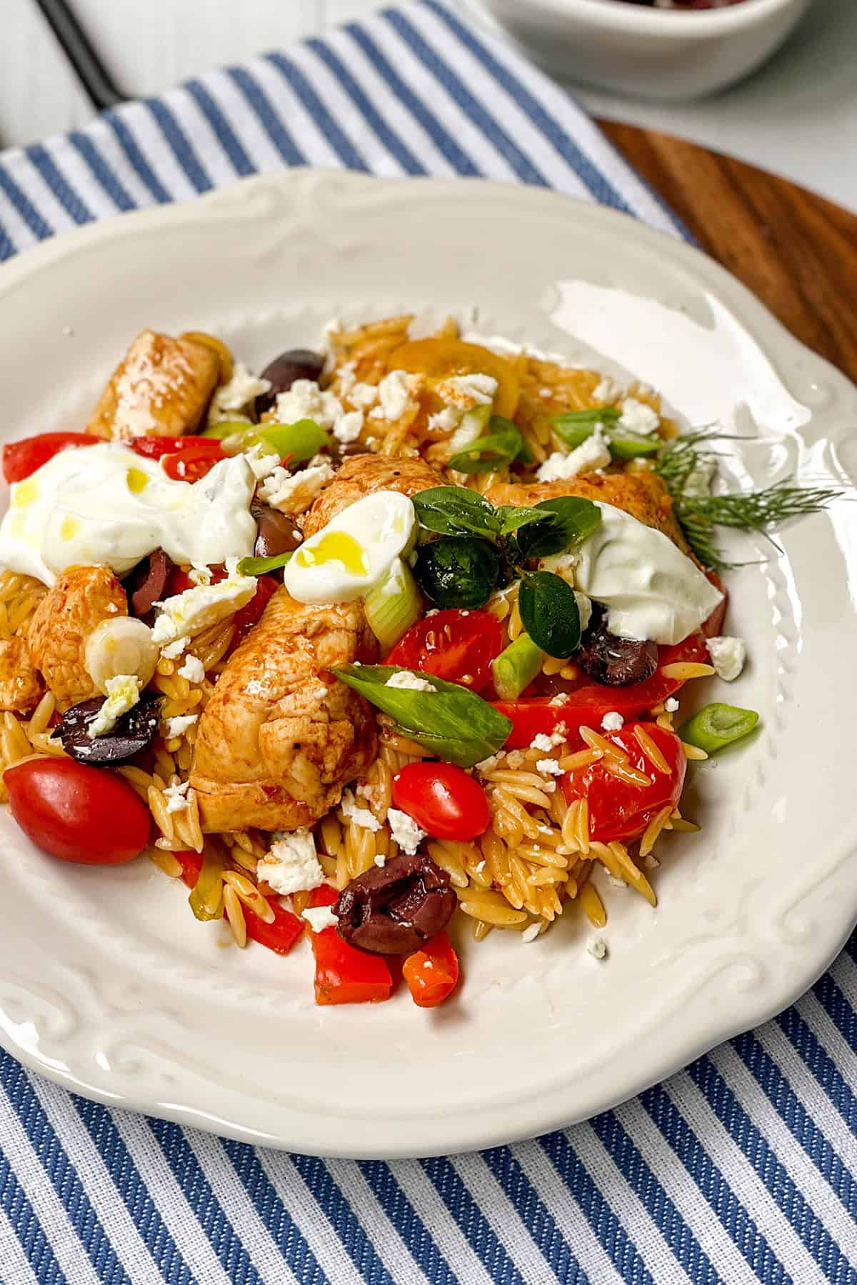 A plate of skillet Greek chicken orzo pasta topped with cherry tomatoes, Kalamata olives, feta, green onions, fresh herbs, and dollops of yogurt sits on a striped napkin.
