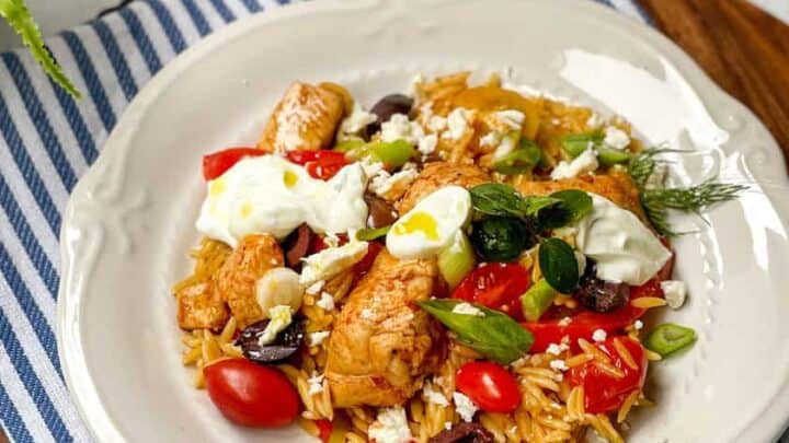 A plate of rice with skillet Greek chicken, cherry tomatoes, olives, feta cheese, and herbs on a white plate, set on a blue-striped napkin with small bowls of toppings in the background.