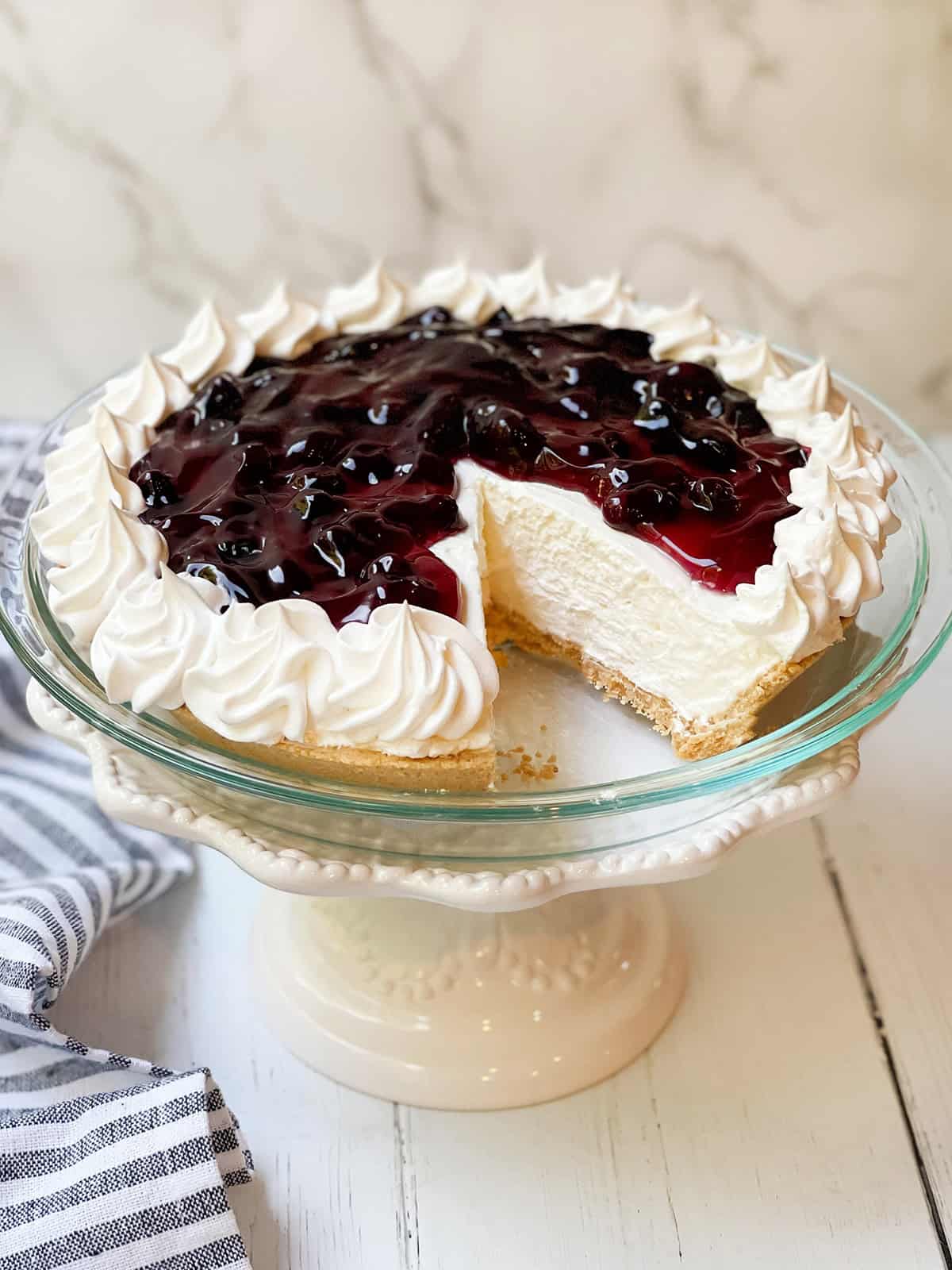 A no bake cheesecake with a whipped cream border and cherry topping sits on a glass cake stand; a slice has been removed, revealing the creamy interior.