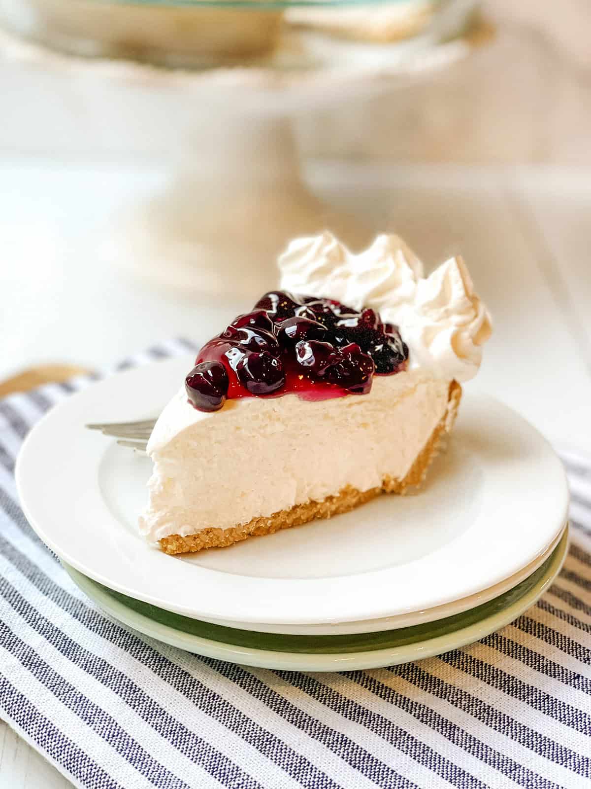 A slice of no bake cheesecake with a graham cracker crust, topped with cherry sauce and whipped cream, sits on a white plate with a fork on a striped napkin.
