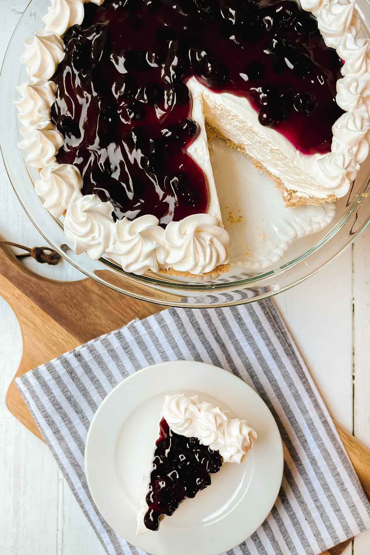 A no bake cheesecake topped with blueberry sauce sits on a glass dish, with one slice served on a small plate beside it.