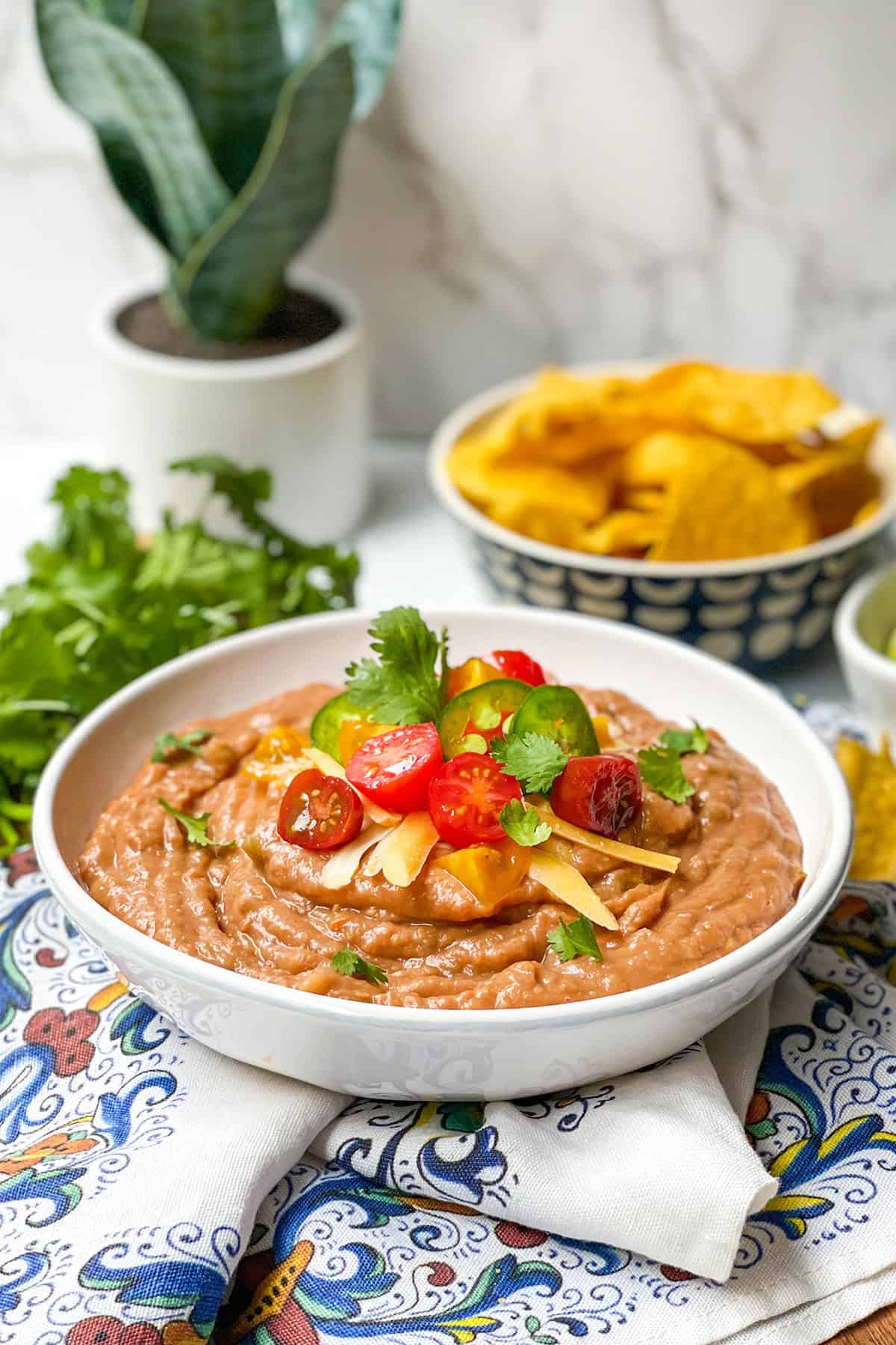 A bowl of refried beans topped with sliced cherry tomatoes, cilantro, and shredded cheese makes a classic bean dip recipe, served with tortilla chips and fresh cilantro in the background.