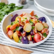 A bowl of German Potato Salad with red, yellow, and purple potatoes, garnished with chopped parsley and pieces of bacon, on a striped cloth.