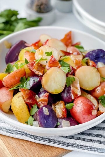 A bowl of German Potato Salad with red, yellow, and purple potatoes, garnished with chopped parsley and pieces of bacon, on a striped cloth.