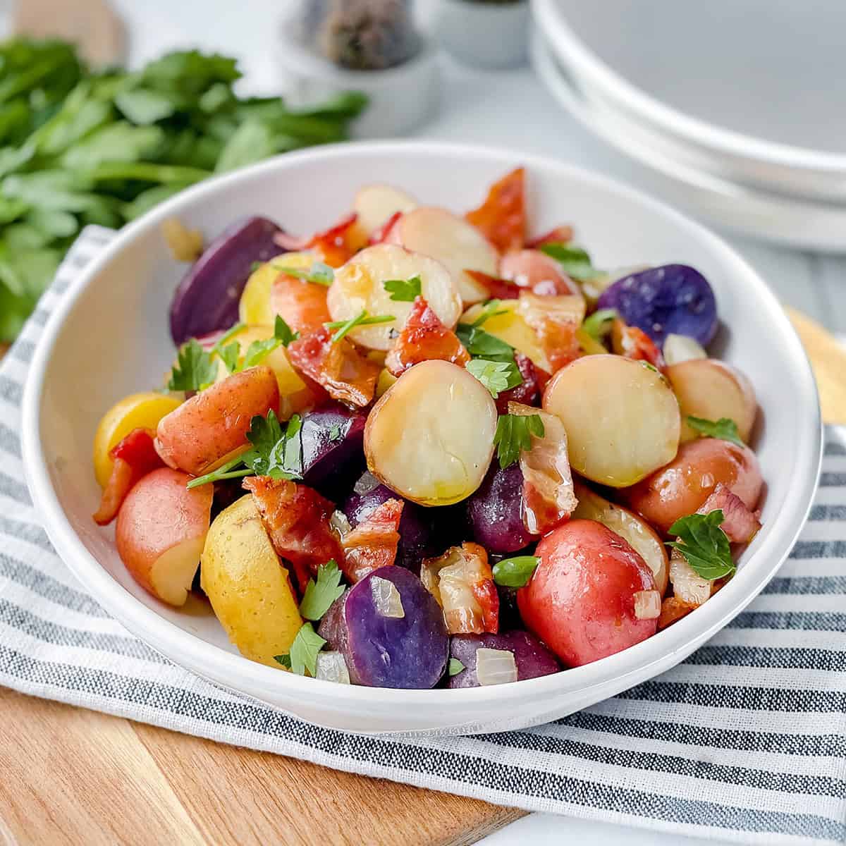 A bowl of German Potato Salad with red, yellow, and purple potatoes, garnished with chopped parsley and pieces of bacon, on a striped cloth.