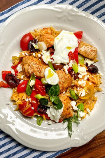 A white plate with orzo pasta, skillet Greek chicken, cherry tomatoes, olives, feta cheese, green onions, and a dollop of yogurt rests on a blue striped cloth.