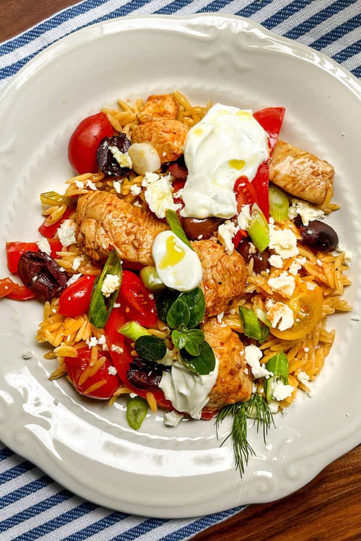 A white plate with orzo pasta, skillet Greek chicken, cherry tomatoes, olives, feta cheese, green onions, and a dollop of yogurt rests on a blue striped cloth.