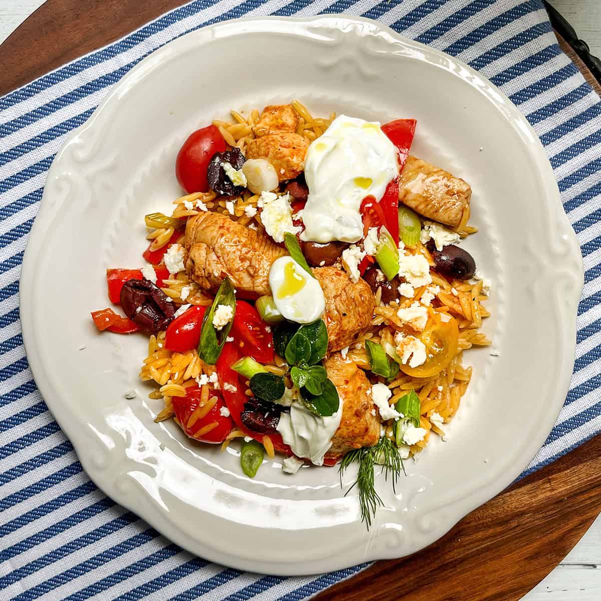 A white plate with orzo pasta, skillet Greek chicken, cherry tomatoes, olives, feta cheese, green onions, and a dollop of yogurt rests on a blue striped cloth.