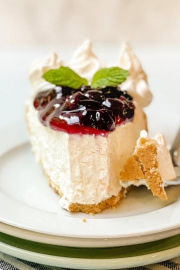 A slice of no bake cheesecake topped with berry sauce and mint leaves, served on a white plate with a fork and a small piece broken off.