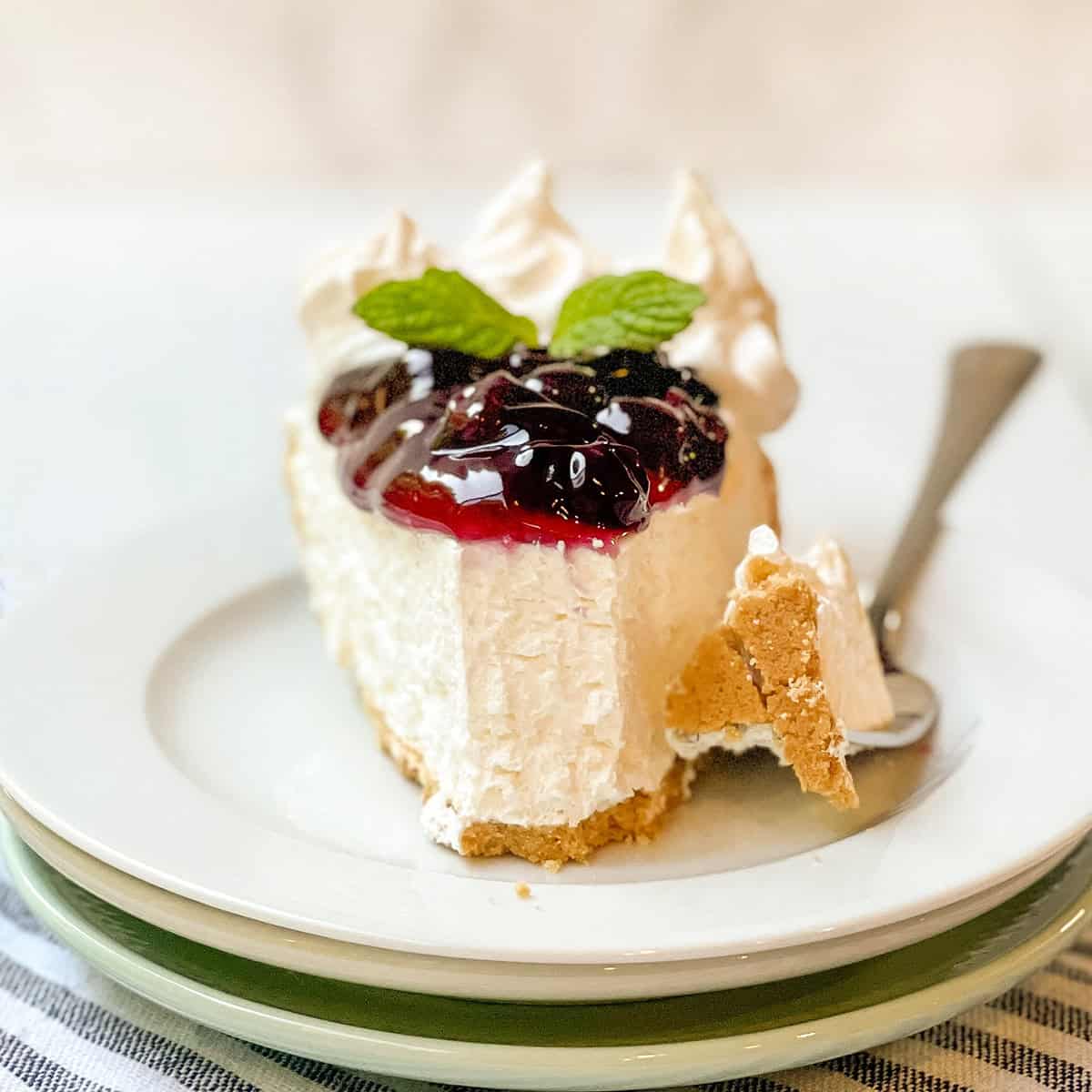 A slice of no bake cheesecake topped with berry sauce and mint leaves, served on a white plate with a fork and a small piece broken off.