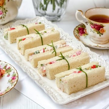 A plate of rectangular strawberry chicken salad tea sandwiches filled with a creamy mixture, each tied with a chive, is served beside floral-patterned teacups filled with tea.