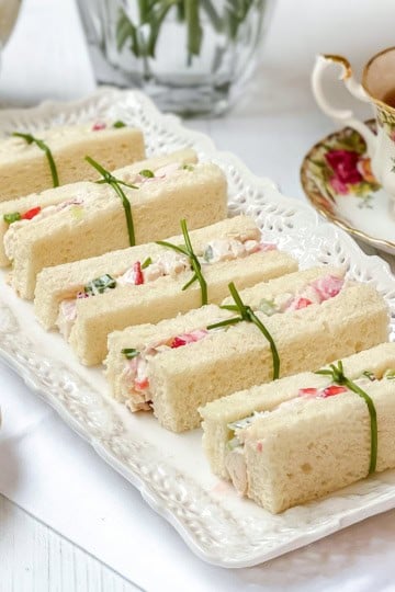 A plate of rectangular strawberry chicken salad tea sandwiches filled with a creamy mixture, each tied with a chive, is served beside floral-patterned teacups filled with tea.