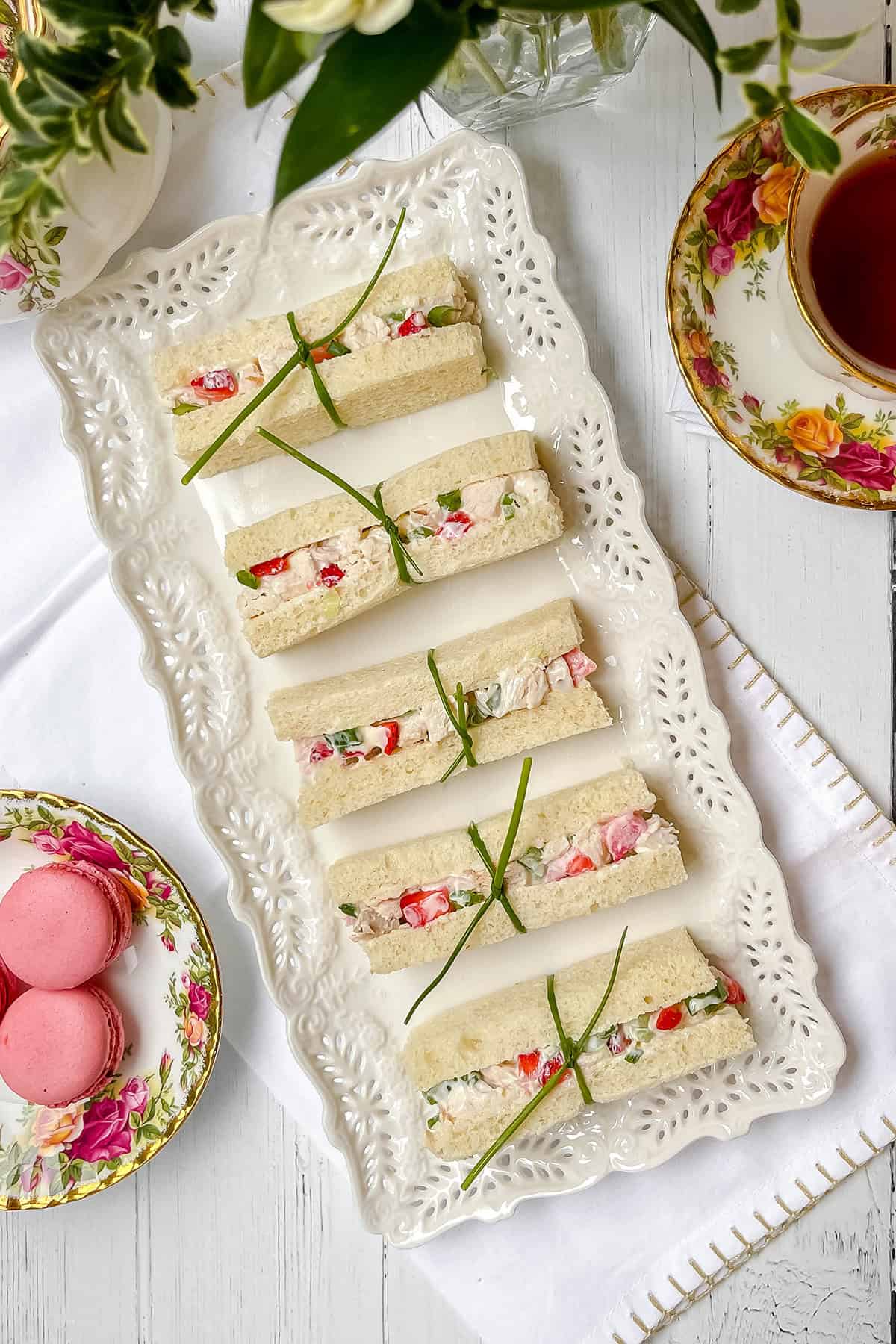 Rectangular strawberry chicken salad tea sandwiches filled with a creamy mixture and vegetables, garnished with chives, are arranged on a decorative white tray beside floral teacups, a teapot, and pink macarons.