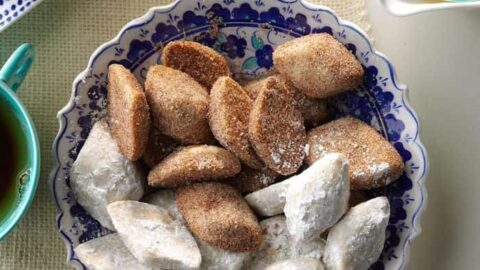 A blue and white patterned plate with powdered sugar-coated and cinnamon-sugar-coated cookies, placed on a woven mat beside a cup of tea—perfect for enjoying after healthy Mexican dinners.