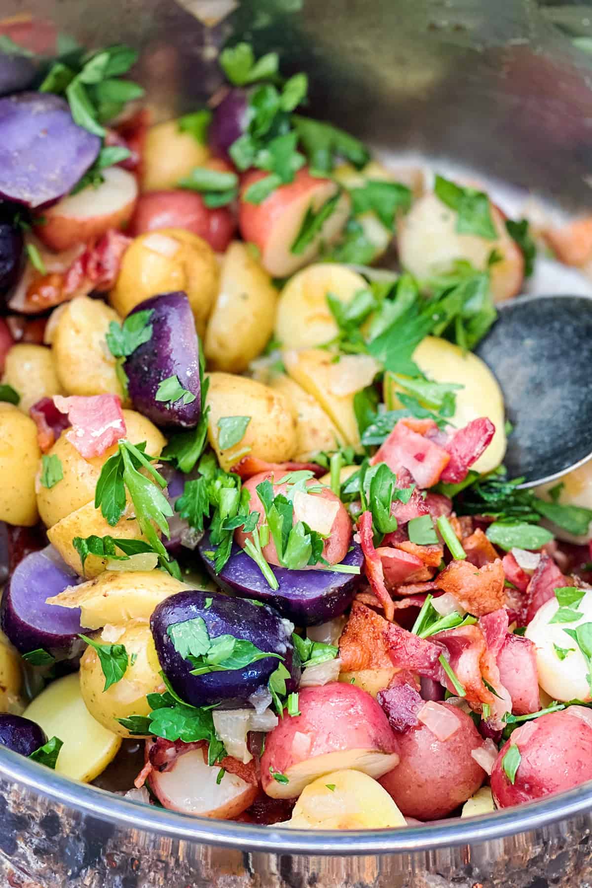 A close-up of German Potato Salad with red, yellow, and purple potatoes, chopped bacon, parsley, and onions, all mixed together in a metal bowl with a serving spoon.