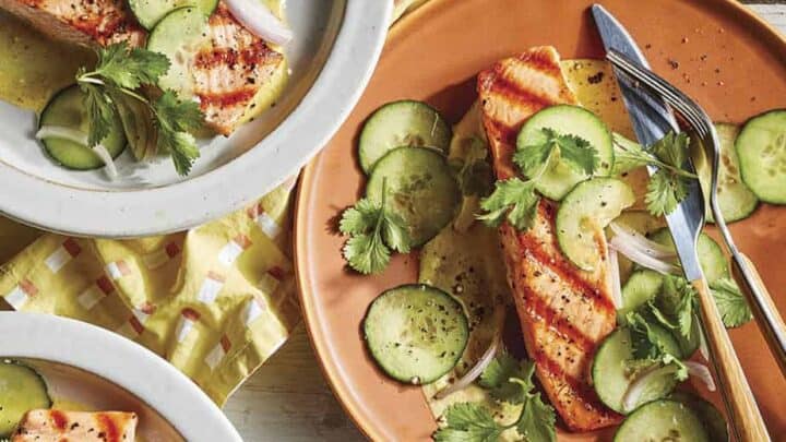 Enjoy healthy May seasonal dinners with grilled salmon fillets served alongside sliced cucumbers, onions, cilantro, and sauce on ceramic plates, complete with napkins and cutlery on a wooden table.