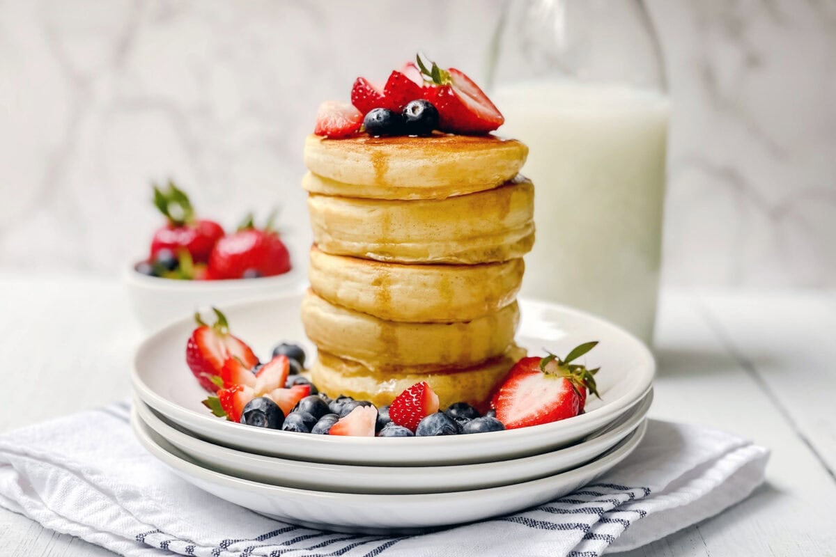 A stack of fluffy pancakes from a classic fluffy pancake recipe is topped with strawberries and blueberries on a white plate, with extra berries scattered around. In the background, a bowl of strawberries and a bottle of milk sit on a white surface.