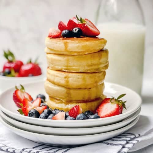 A stack of fluffy pancakes from a classic fluffy pancake recipe is topped with strawberries and blueberries on a white plate, with extra berries scattered around. In the background, a bowl of strawberries and a bottle of milk sit on a white surface.