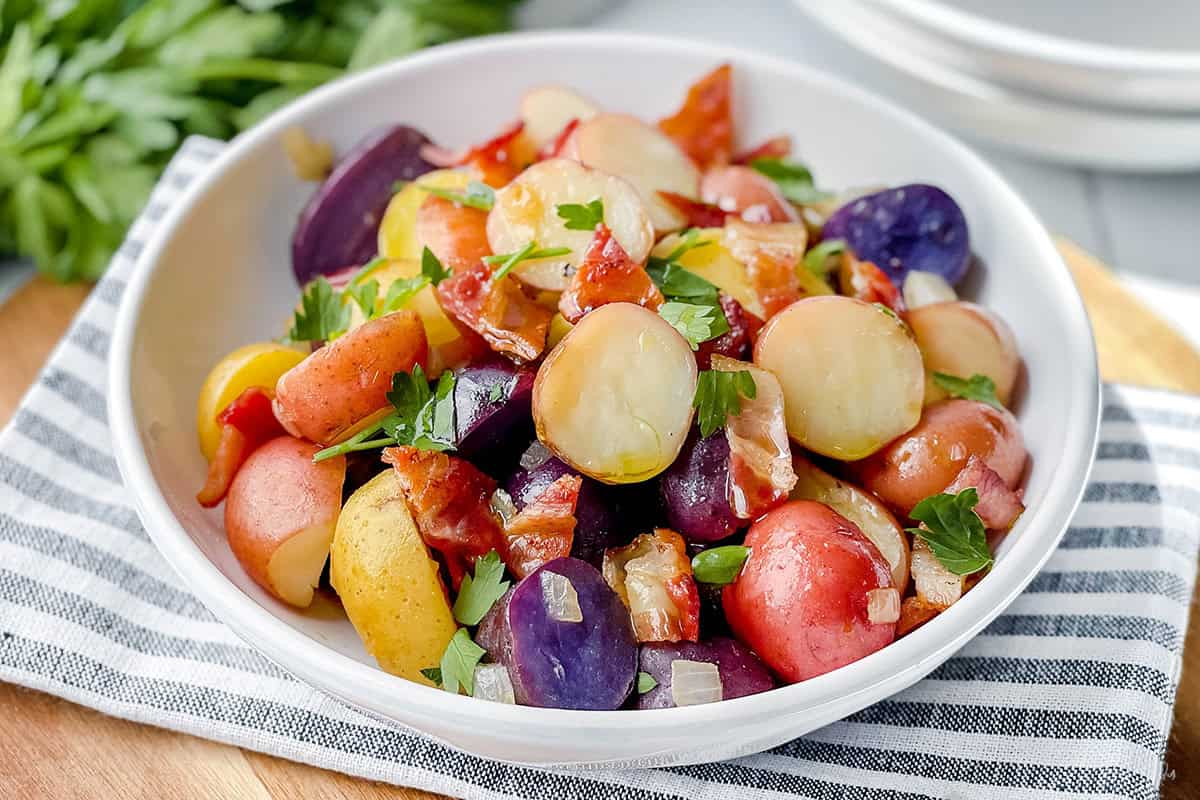 A bowl of mixed roasted potatoes, including purple, red, and yellow varieties, inspired by German Potato Salad, garnished with parsley and pieces of bacon.
