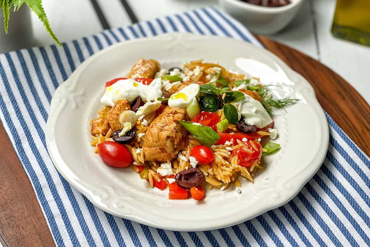 A plate of skillet Greek chicken with orzo, cherry tomatoes, kalamata olives, feta cheese, and dollops of yogurt on a white dish, set on a striped cloth.