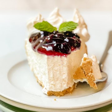 A slice of no bake cheesecake with a graham cracker crust, topped with berry sauce, whipped cream, and mint leaves on a white plate with a fork.