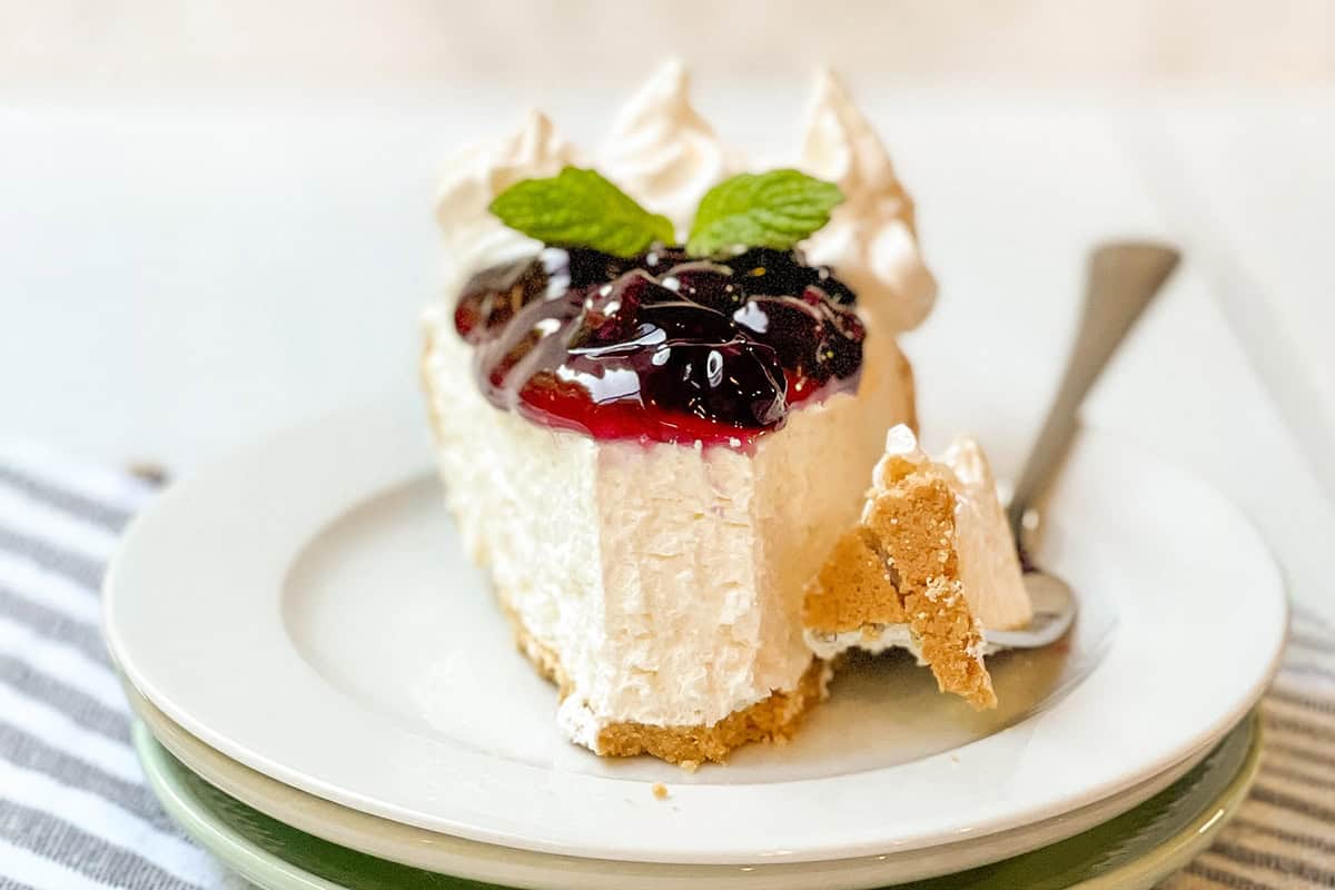 A slice of no bake cheesecake with a graham cracker crust, topped with berry sauce, whipped cream, and mint leaves on a white plate with a fork.