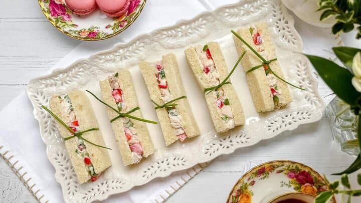 Six rectangular strawberry chicken salad tea sandwiches garnished with chives are arranged on a decorative white platter, accompanied by a plate of pink macarons and floral china teacups.