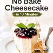 A slice of no bake cheesecake topped with blueberry sauce and mint, served on a white plate with a fork, and accompanied by a dollop of whipped cream.