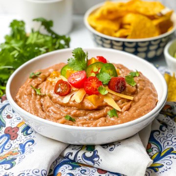 A bowl of refried beans topped with sliced cherry tomatoes, cheese, and cilantro makes this the perfect Bean Dip Recipe, served with crispy tortilla chips and fresh cilantro in the background.