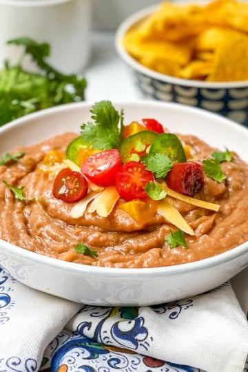 A bowl of refried beans topped with sliced cherry tomatoes, cheese, and cilantro makes this the perfect Bean Dip Recipe, served with crispy tortilla chips and fresh cilantro in the background.