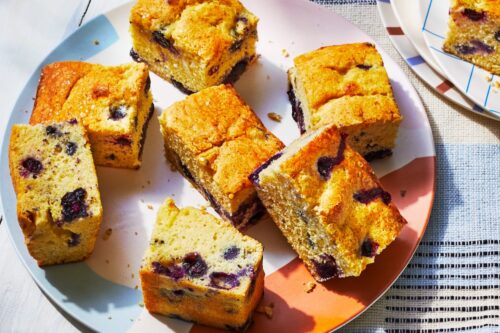 A plate with eight pieces of blueberry cornbread, perfect for picnic desserts, is displayed on a patterned tablecloth, with a separate plate holding one more piece in the background.