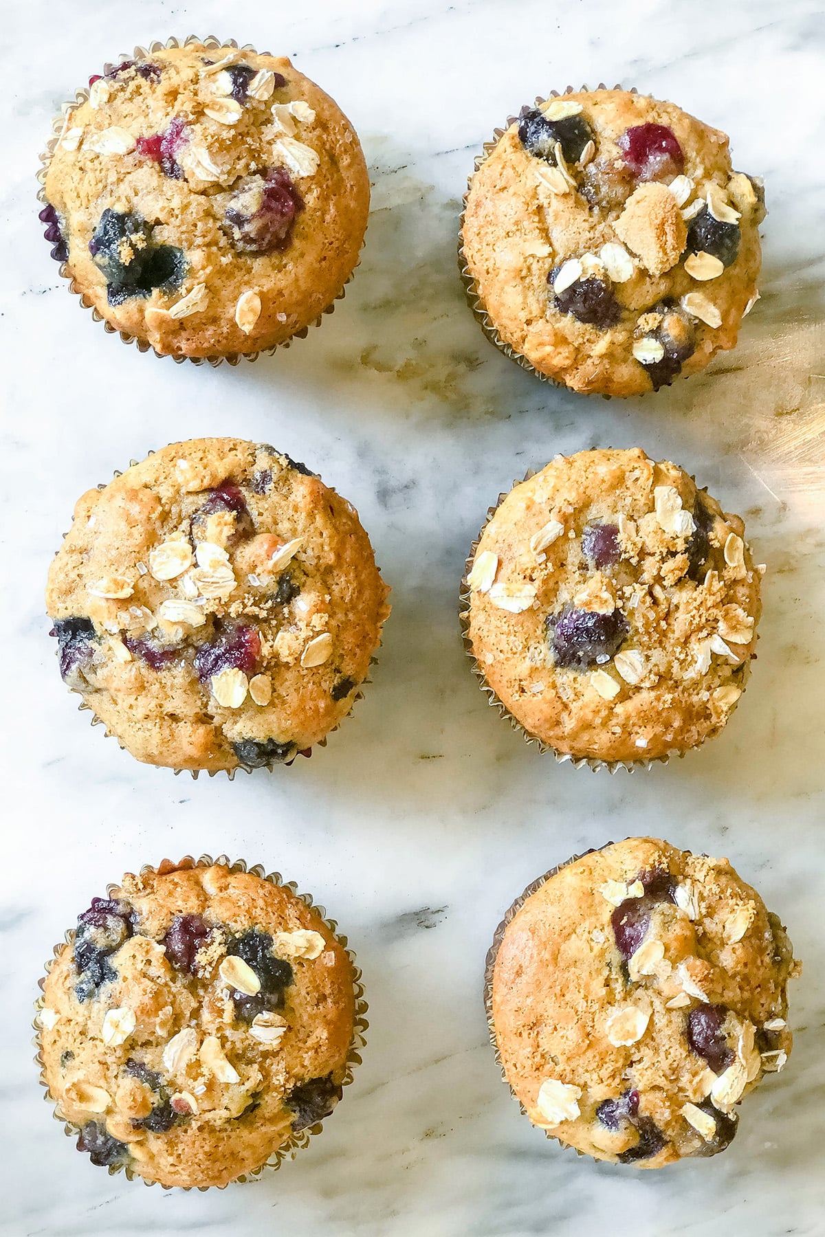 Six blueberry oat muffins with oats on top are arranged in two rows on a white marble surface.