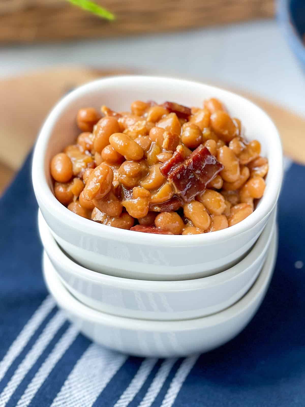 A white bowl filled with Boston baked beans and pieces of bacon, stacked on two empty bowls, sits on a blue and white striped cloth.