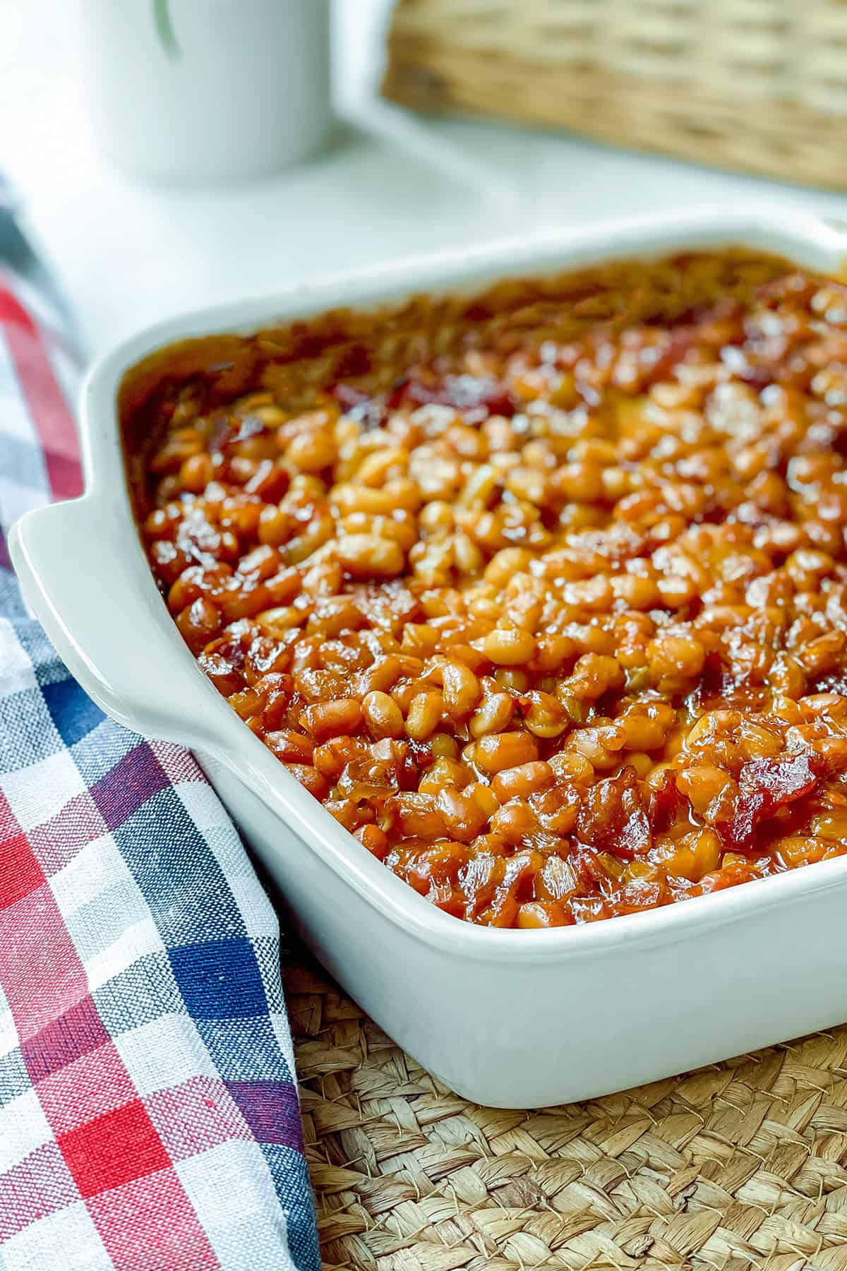 A close-up of Boston baked beans in a white square baking dish, with a red, white, and blue plaid cloth beside it on a woven surface.