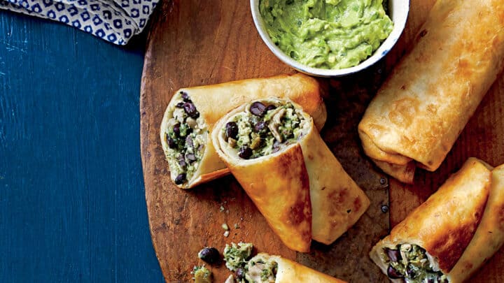 Crispy chimichangas filled with beans and vegetables are arranged on a wooden board, served with guacamole and a tomato salad—a perfect addition to your July 20 Minute Dinners lineup.