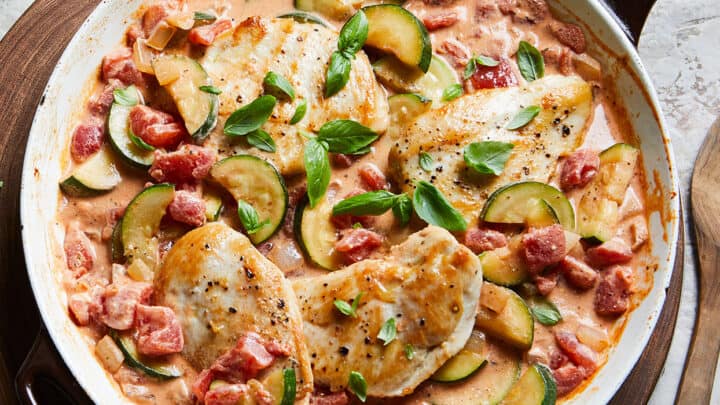 A skillet with cooked chicken breasts in a creamy tomato sauce, accompanied by sliced zucchini and diced tomatoes, garnished with fresh basil leaves&mdash;a perfect choice for 20 Minute Skillet Dinners.