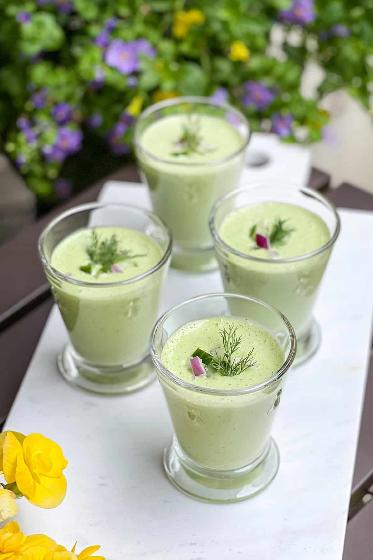 Four glass cups filled with pale green cucumber soup, garnished with dill and small pieces of red onion, placed on a white tray with flowers in the background.