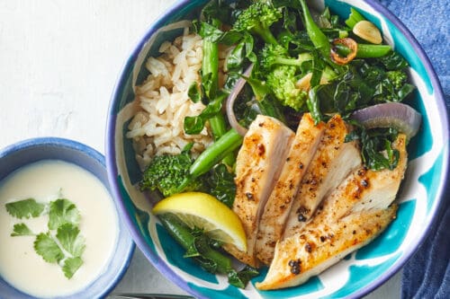 A bowl with grilled chicken, brown rice, greens, and lemon wedge next to a small bowl of creamy sauce with cilantro garnish-perfect for high protein summer dinners-served with a fork and blue napkin on a white table.