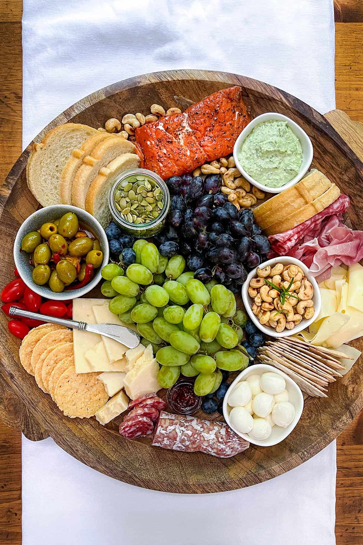 A Mediterranean charcuterie board features a wooden platter with grapes, olives, cheeses, crackers, bread, cured meats, nuts, smoked salmon, and dips arranged in sections on a white cloth.