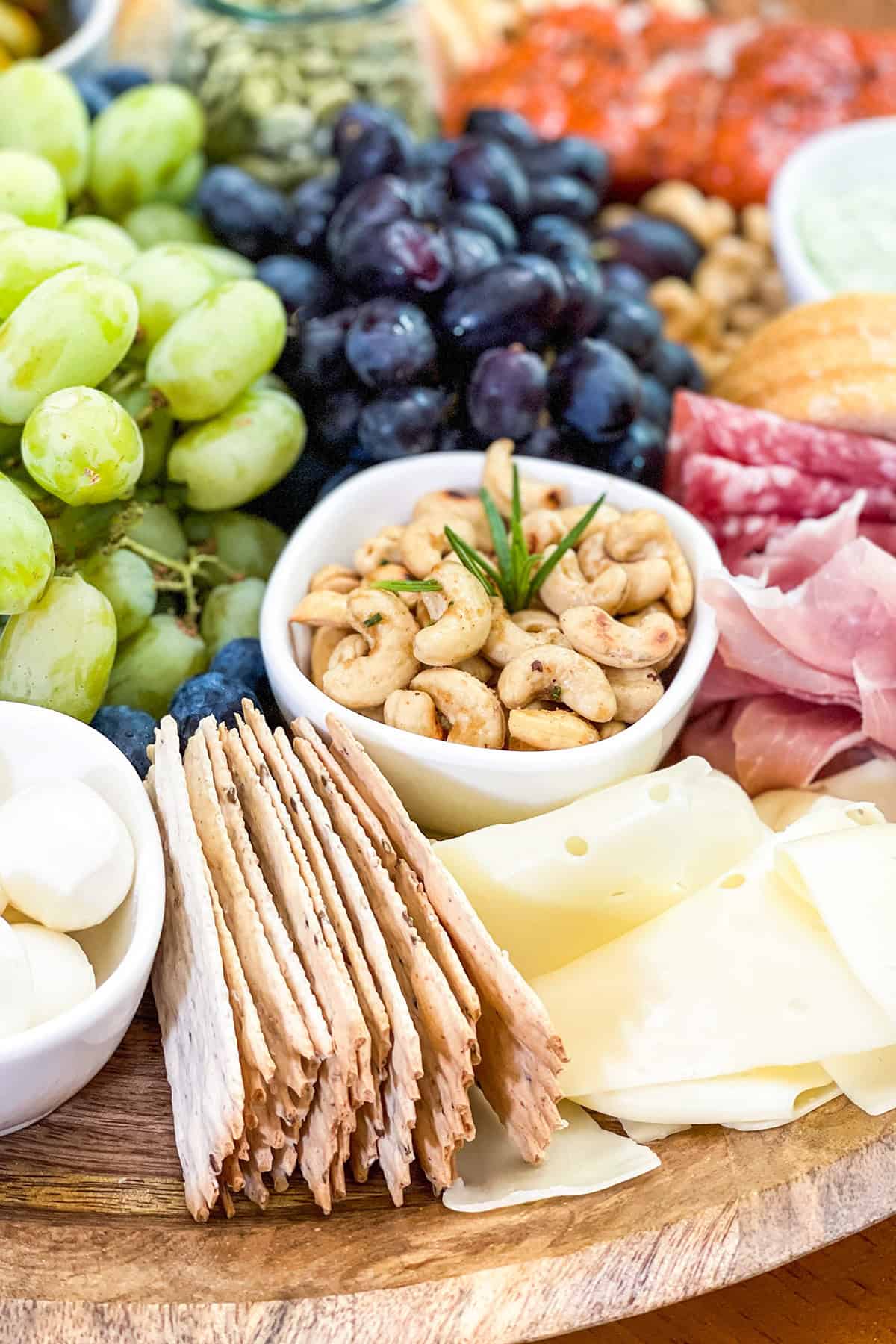A Mediterranean charcuterie board features a wooden platter with crackers, sliced cheese, mozzarella balls, seasoned cashews, green and black grapes, salami, and prosciutto.