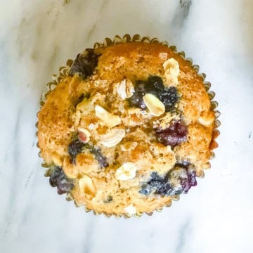 A single blueberry oat muffin with oats on top, in a paper liner, sits on a white marble surface.